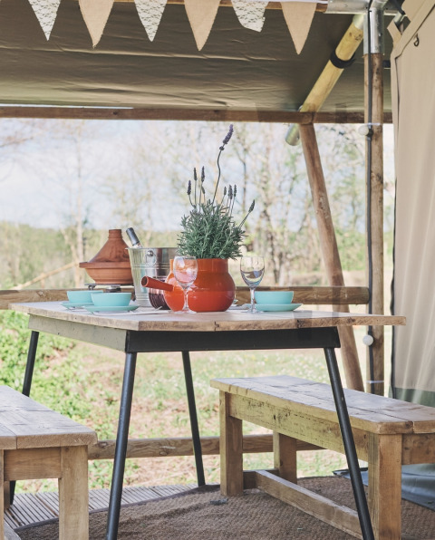 Table de pique-nique sous tente safari avec bancs en bois, vaisselle et pot de lavande décoratif.