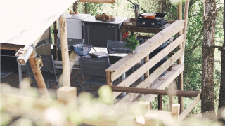 Vue latérale d’une terrasse de tente safari avec cuisine extérieure et coin salon au milieu des arbres.