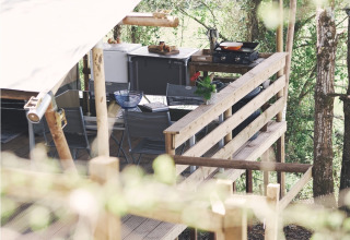 Vue latérale d’une terrasse de tente safari avec cuisine extérieure et coin salon au milieu des arbres.