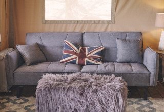 Interior de tienda safari en Camping d'Artagnan en Francia con sofá gris, cojín británico y banco peludo.
