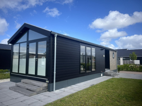 Chalet moderne appelé Sea Pearl avec de grandes fenêtres, façade noire et terrasse pavée sous ciel bleu.