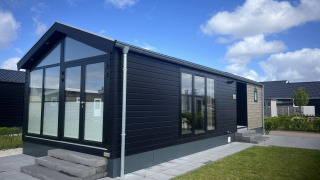 Moderne chalet ved navn Sea Pearl med store vinduer, sort facade og flisebelagt terrasse under blå himmel.