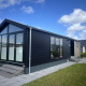 Moderne chalet ved navn Sea Pearl med store vinduer, sort facade og flisebelagt terrasse under blå himmel.