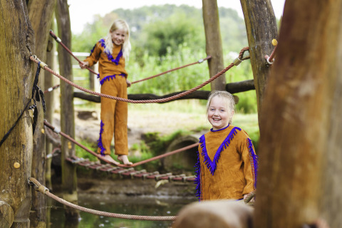 Zwei Kinder in bunten Kostümen spielen auf einer Seilbrücke im Beloofde Land Ferienpark in Gelderland.