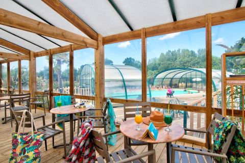 Terraza cubierta con mesas y bebidas coloridas, vista a la piscina interior de un parque vacacional francés.