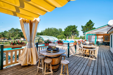 Sonnige Terrasse mit Fass-Tischen und Holzstühlen auf Camping Paradis La Bretonniere, Pays de la Loire, Frankreich.