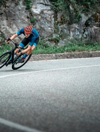 Ciclista con casco rojo toma una curva cerrada en carretera asfaltada junto a rocas y vegetación.