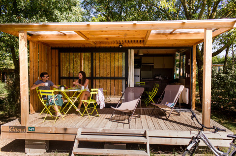 Dos personas desayunan en la terraza de madera de una cabaña en Huttopia Saumur, Pays de la Loire, Francia.