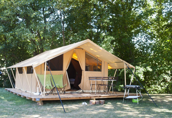 Safari-telt på træterrasse i en skovlysning med stole, bord og grill under sommersol.
