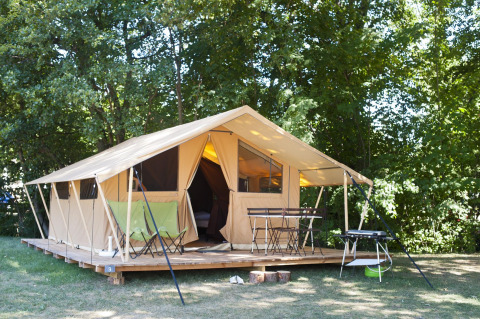 Safaritent op houten vlonder in een groene omgeving met stoelen, tafel en barbecue in het zonlicht.