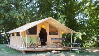 Safaritent op houten terras in bosrijke omgeving met campingstoelen, tafel en barbecue in de zon.