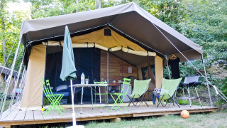 Safari tent Toile&Bois Sweet met houten terras, stoelen, tafel en barbecue omgeven door bomen.