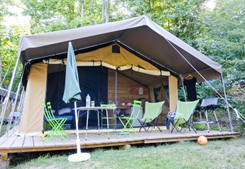 Tente safari Toile&Bois Sweet avec terrasse en bois, chaises, table et barbecue, entourée d’arbres.
