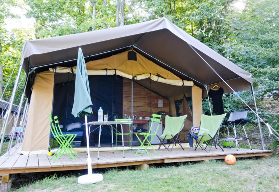 Safari tent Toile&Bois Sweet met houten terras, stoelen, tafel en barbecue omgeven door bomen.