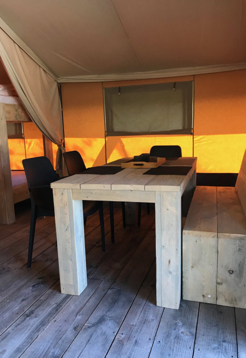 Vista interior de una tienda safari en Camping Paradis La Bretonniere con mesa y sillas de madera.
