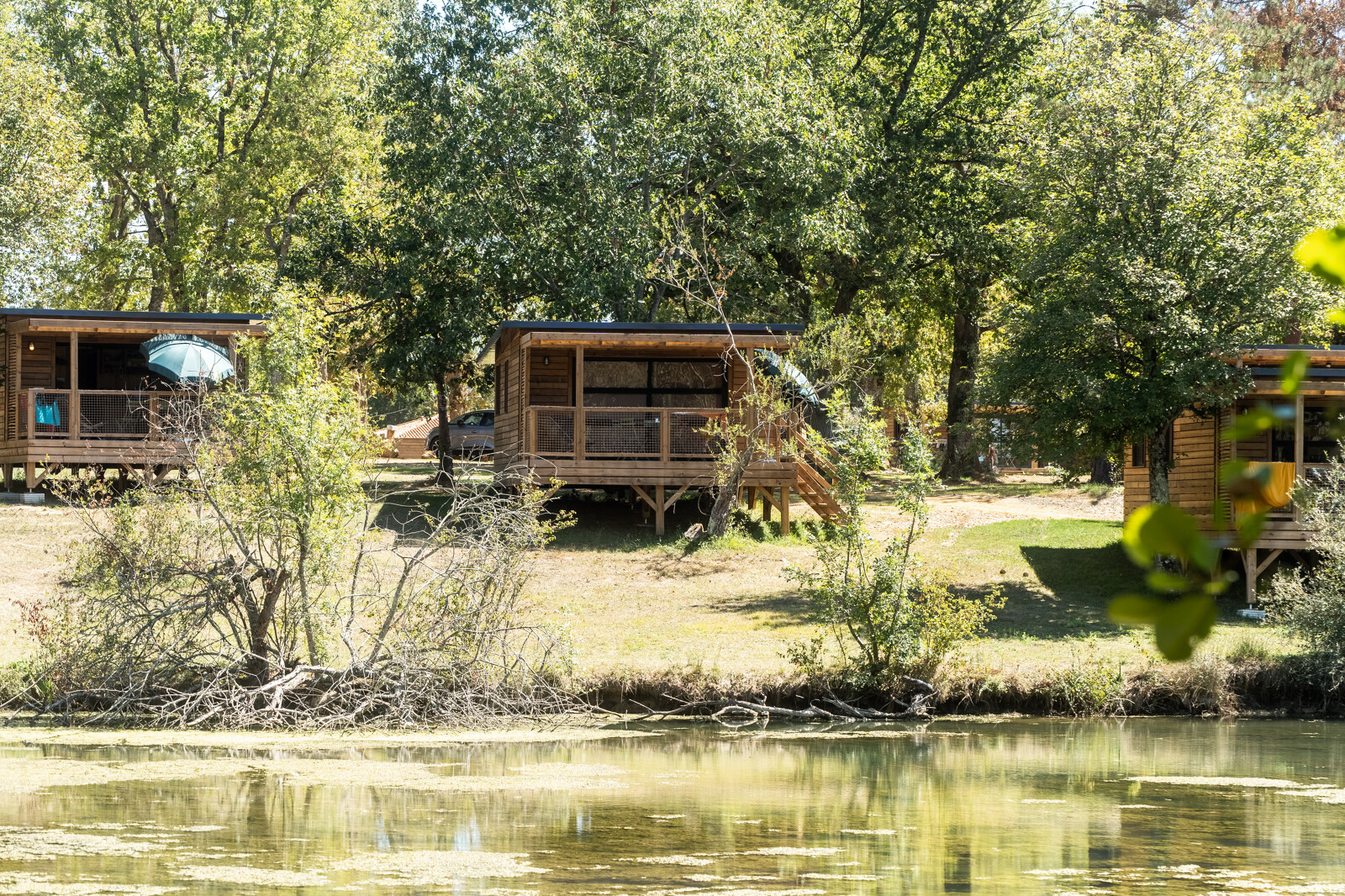 Houten chalets aan het meer bij Chalet Evasion, Huttopia Lac de l'Uby - Gers, omgeven door bomen.
