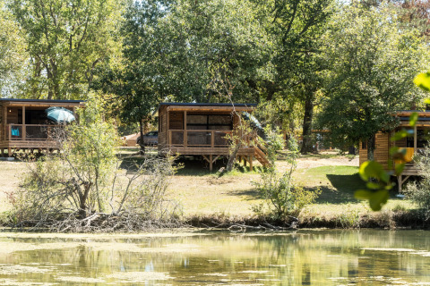 Træhytter ved søbredden i Chalet Evasion, Huttopia Lac de l'Uby - Gers, omgivet af frodige træer.
