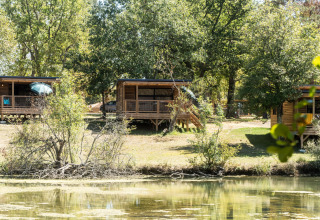 Cabañas de madera junto al lago en Chalet Evasion, Huttopia Lac de l'Uby - Gers, rodeadas de árboles.