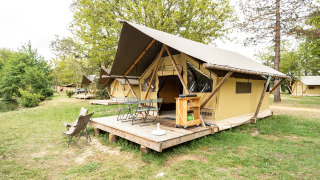 Tenda safari Trappeur con veranda in legno, sedie da esterno e alberi in un'area campeggio verde.