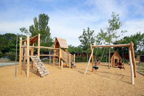 Parque infantil de madera con columpios en Camping d'Angers - Lac de Maine, niños juegan bajo el sol.
