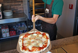 Een lachende pizzabakker op Camping d'Angers - Lac de Maine serveert een vers gebakken pizza aan gasten.