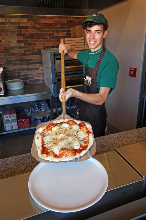 A smiling pizza chef at Camping d'Angers - Lac de Maine serves a freshly baked pizza to guests.