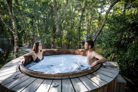 Dos personas se relajan en un jacuzzi al aire libre rodeado de árboles en Village Huttopia Lac de Rillé, Francia.