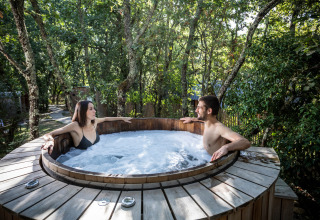 Dos personas se relajan en un jacuzzi al aire libre rodeado de árboles en Village Huttopia Lac de Rillé, Francia.