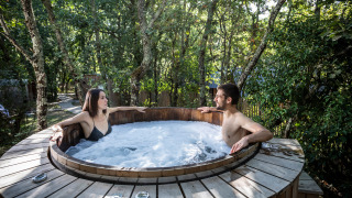 Dos personas se relajan en un jacuzzi al aire libre rodeado de árboles en Village Huttopia Lac de Rillé, Francia.