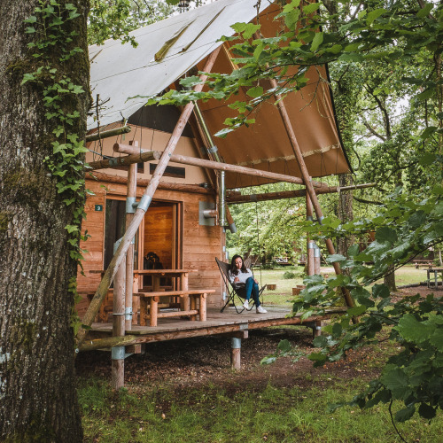 Woman relaxing on the porch of a wooden cabin in the woods at Village Huttopia Lac de Rillé, France.