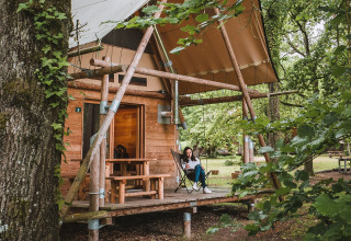 Frau entspannt sich auf der Veranda einer Holzhütte im Wald im Village Huttopia Lac de Rillé, Frankreich.