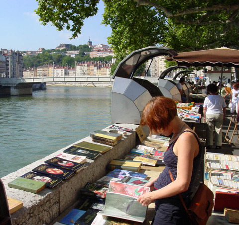 Mujer mirando libros en un mercado al aire libre junto al río en Lyon, Francia, con puente y ciudad detrás.