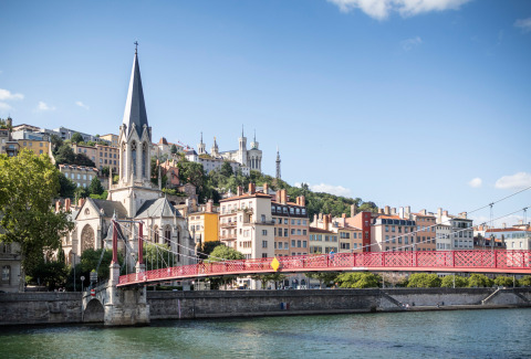 Vista de un puente rojo, una iglesia y edificios coloridos junto al río en Lyon, Francia, en un día soleado.