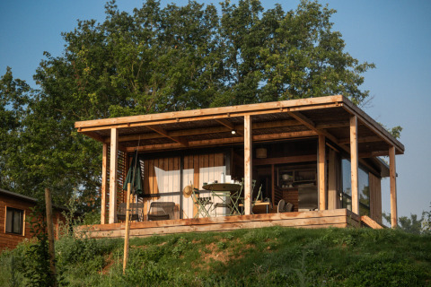 En træhytte med overdækket terrasse omgivet af grønne træer ved Chalet Evasion Vista, Huttopia Pays de Cordes sur Ciel.