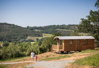 En kvinde og et barn går mod en træhytte med bakker og grønne marker i baggrunden i Occitanie, Frankrig.