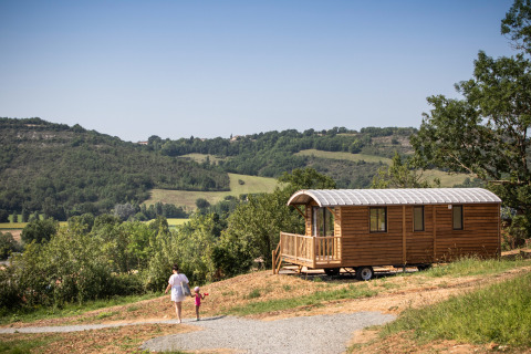 Eine Frau und ein Kind gehen zu einer Holzhütte mit Blick auf grüne Hügel im Ferienpark in Occitanie, Frankreich.
