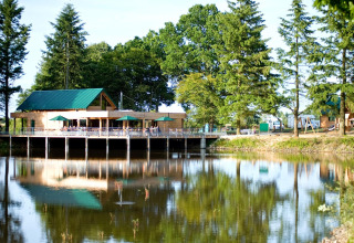 Bâtiment en bois au bord d’un lac avec parasols verts, entouré d’arbres à Village Huttopia Senonches.