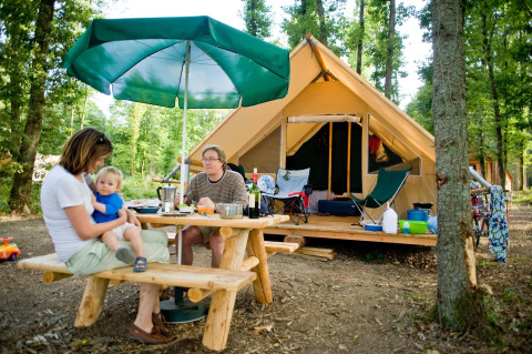 Family enjoying outdoor meal at picnic table with large tent in the forest at Village Huttopia Senonches, France.