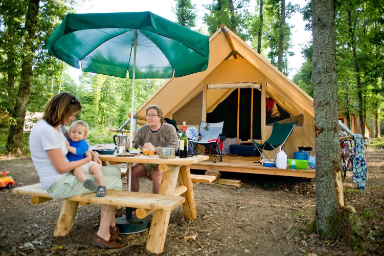 Family enjoying outdoor meal at picnic table with large tent in the forest at Village Huttopia Senonches, France.