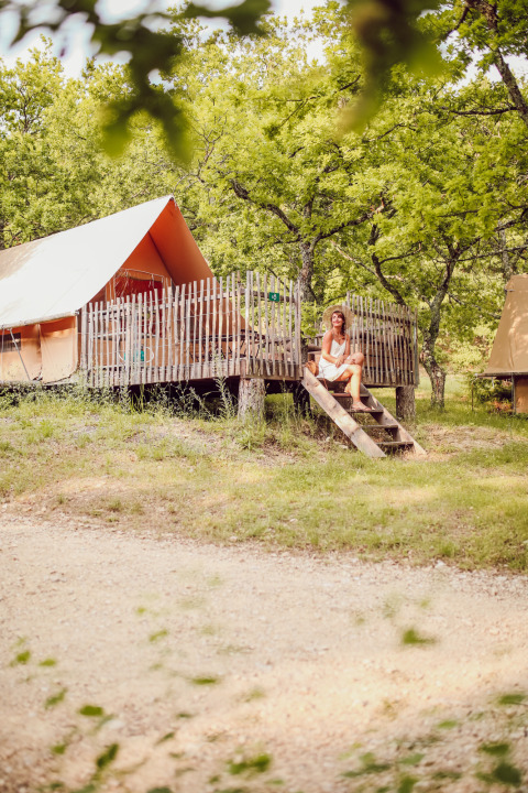 Woman sits on the steps of a glamping tent surrounded by trees at Village Huttopia Dieulefit in France.