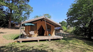 Tienda safari Tent Sweet con terraza, sillas y sombrilla, rodeada de naturaleza bajo un cielo azul claro.