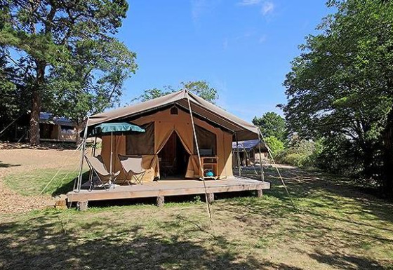 Tienda safari Tent Sweet con terraza, sillas y sombrilla, rodeada de naturaleza bajo un cielo azul claro.