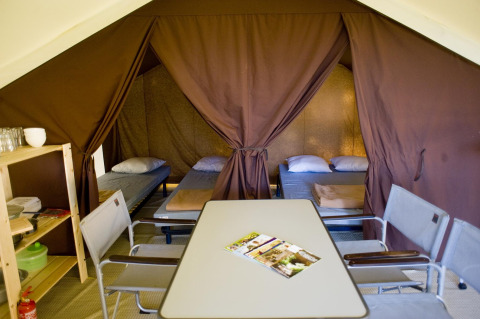 Interior de una tienda safari bungalow CLASSIC IV con camas, mesa y sillas en Huttopia Lac d’Aiguebelette, Francia.
