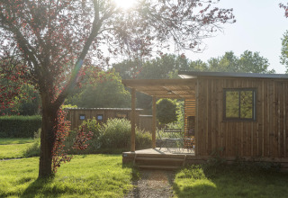 Cabaña de madera soleada con porche rodeada de árboles y césped en Camping Valkenburg, Países Bajos.