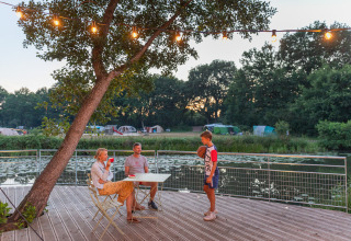 Familie nyder aftenen ved et bord på en terrasse med udsigt til sø og telte på Huttopia De Roos, Overijssel.