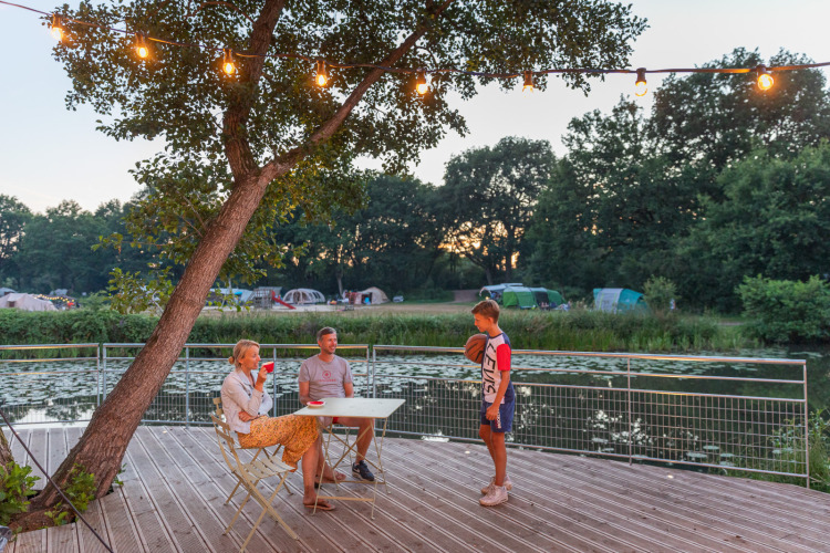 Family enjoys evening on deck overlooking a lake and tents at Huttopia De Roos holiday park, Overijssel, Netherlands.