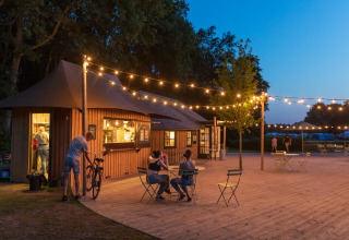 Udendørs caféområde ved skumring i Huttopia De Roos med lyskæder og træbygninger i Overijssel.