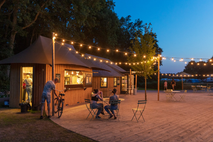 Outdoor café with string lights and wooden cabins at Huttopia De Roos holiday park, Overijssel, Netherlands.