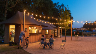 Cafetería al aire libre con luces y cabañas de madera en Huttopia De Roos, Overijssel, Países Bajos.