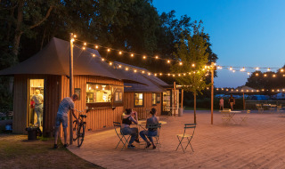 Cafetería al aire libre con luces y cabañas de madera en Huttopia De Roos, Overijssel, Países Bajos.